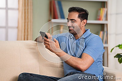 Happy relaxed indian man using mobile phone while sitting on sofa at home during sunday - concept of online dating Stock Photo