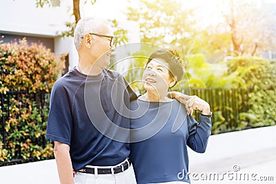 Happy retired senior Asian couple walking and looking at each other with romance in outdoor park and house in background. Stock Photo