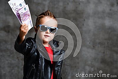 Happy rich kid boy millionaire in fashion sunglasses hold stack of 500 euro money cash in leather jacket and red t-shirt Stock Photo