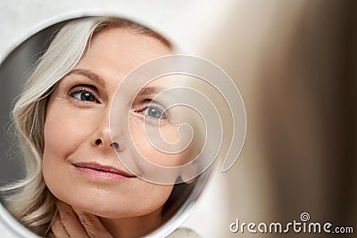 Happy 50s middle aged woman touching face skin looking in mirror reflection. Stock Photo