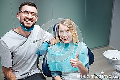Happy satisfied young woman sit in char and smile. She hold big thumb up. Male dentist stand beside her and smile too Stock Photo