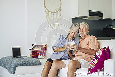 Happy senior adult couple of cacuasian people in love sitting at home on the sofa and taking a wood hand made hearth on hands. Stock Photo