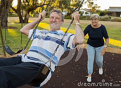 Happy senior American couple around 70 years old enjoying at swing park with wife pushing husband smiling and having fun together Stock Photo