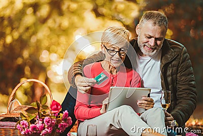 Senior couple having a picnic in autumn park using digital tablet and credit card for online shopping Stock Photo