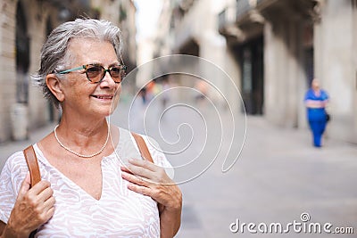 Happy senior woman walking in the city pedestrian street wearing sunglasses and backpack. Caucasian gray haired woman Stock Photo