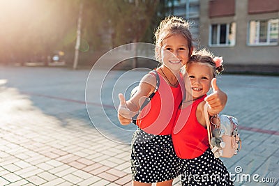 Happy sisters girls wearing backpacks and showing thumbs up. Kids pupils smiling outdoors school building. Education Stock Photo