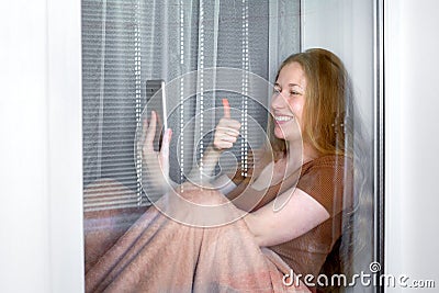 Happy smile girl sits on windowsill by window and holding smartphone. Stock Photo