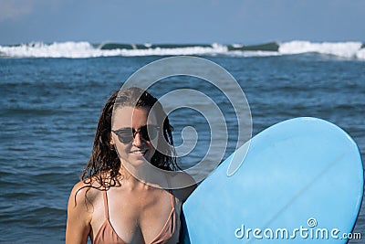 Happy smile young brunette woman hold blue surfboard going out of ocean after morning surfing. Portrait of beautiful girl spending Stock Photo