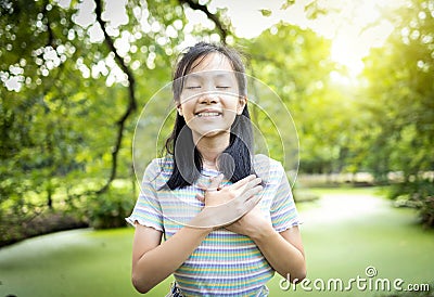 Happy smiling asian child girl standing in green nature,hold hands on heart,feel peace of mind,female teenage closed eyes,enjoy Stock Photo