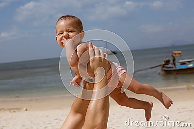 Happy smiling baby in dad arms. On a tropical beach. Sunny day, father throws up infant toddler, hold on hands. Child enjoyed Stock Photo