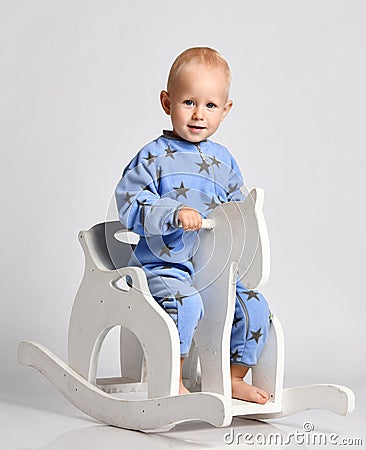 Happy smiling barefooted baby boy in blue fleece jumpsuit with stars plays rides white kids rocking horse toy Stock Photo