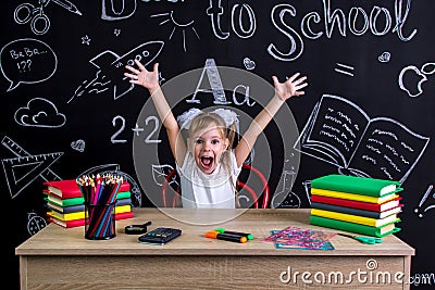 Happy, smiling and excited schoolgirl sitting at the desk with both arms up, surrounded with school supplies. Chalkboard Stock Photo