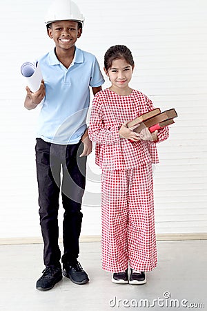 Happy smiling kid engineer African boy with helmet safety hard hat holding blueprint and cute Asian girl holding pile of books Stock Photo