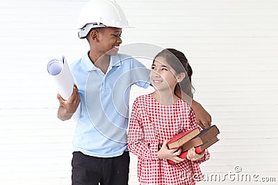 Happy smiling kid engineer African boy with helmet safety hat holding blueprint and cute Asian girl holding pile books standing Stock Photo