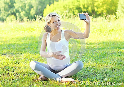 Happy smiling pregnant woman on grass and makes self-portrait on smartphone Stock Photo