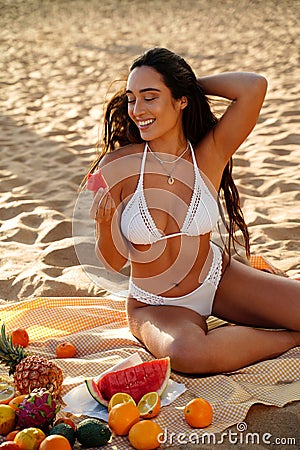Happy, smiling woman in white bikini eating fruits, sitting at the sandy beach, enjoying sunset time, relaxing. Island girl Stock Photo