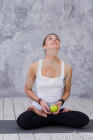 Happy sports woman holding apple sitting on a yoga mat over gray background Stock Photo