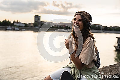 Happy teenage female tourist sitting on sea promenade of an old city in sunset. Young woman world traveler singing her favorite Stock Photo