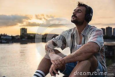 Happy teenage male tourist sitting on sea promenade of an old city in sunset. Young man world traveler singing his favorite song Stock Photo