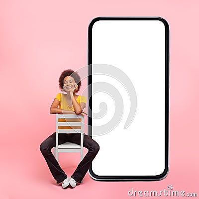 Happy thinking young mixed race lady with curly hair in casual sits on chair near huge smartphone with empty screen Stock Photo
