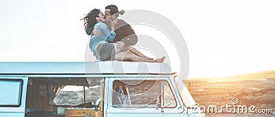 Happy travel influencers couple kissing on minivan roof at sunset with desert on background - Trendy people having fun traveling Stock Photo