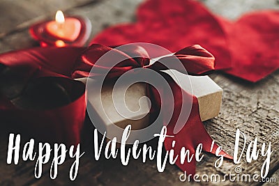 Happy Valentine`s day text on stylish gift box with red ribbon and red velvet hearts on rustic wood Stock Photo