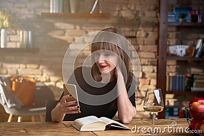 Happy white woman reading book, using phone, checking social media newsfeed or e-mails at home in the living room, drinking wine. Stock Photo