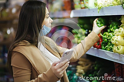 Happy woman in face mask hold phone and choose vegetables in supermarket. Beautiful young business woman choosing Stock Photo