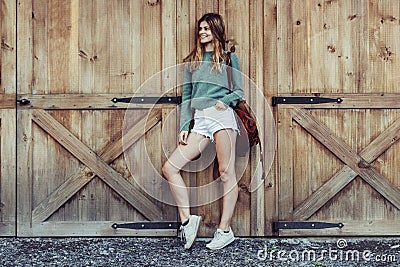 Happy woman with long legs look to the side near barn on the farm wearing casual outfit with shorts, backpack and sneakers. Stock Photo