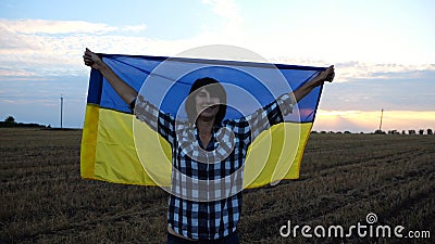 Happy woman looks into camera standing on wheat field with a lifted blue-yellow banner at sunset. Ukrainian smiling lady Stock Photo