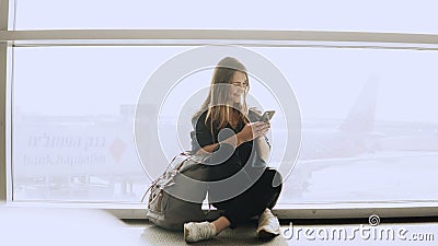 Happy woman sits with smartphone by airport window. Caucasian girl with backpack using messenger app in terminal. 4K. Stock Photo