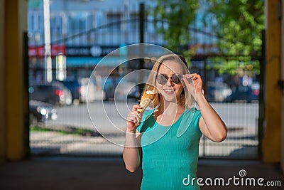 A happy woman in a turquoise dress stands in the yard and eats a waffle cone on a warm summer day. Beautiful blonde in sunglasses Stock Photo
