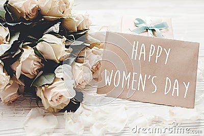 Happy Women`s Day text sign on craft greeting card and white roses bouquet, gift box on wooden background. International womens Stock Photo