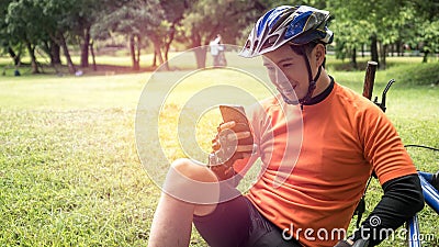 Happy young asian male cyclist in orange sportswear using Mobile Phone In Park, Beautiful sunny day Stock Photo