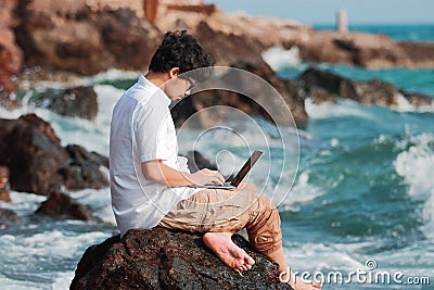 Happy young Asian man with computer laptop sitting on the rock of seashore in summer time. Summer vacations and lifestyle concept Stock Photo