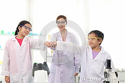 Happy young Asian student schoolgirls in lab coat with female scientist teacher bumping their fists together to celebrate success Stock Photo