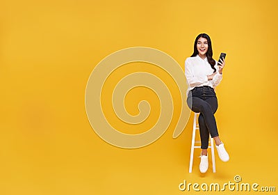 Happy young Asian woman showing mobile phone. While her sitting on white chair and looking on yellow copy space background Stock Photo