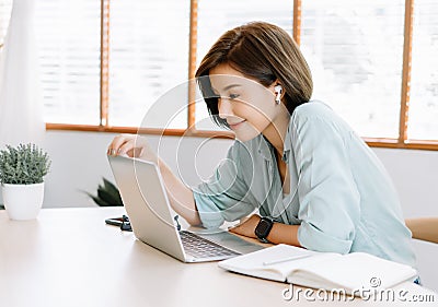 Happy young beautiful Asian woman using laptop working remote and wireless earphones sitting at table in home office.Remote work,E Stock Photo
