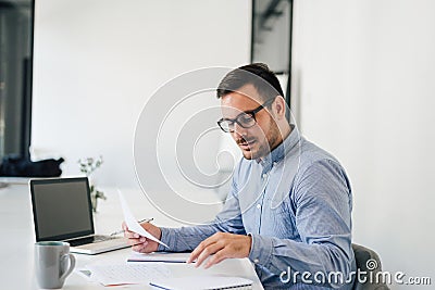 Happy young businessman looking at papers graphs and charts using laptop at his office desk Stock Photo