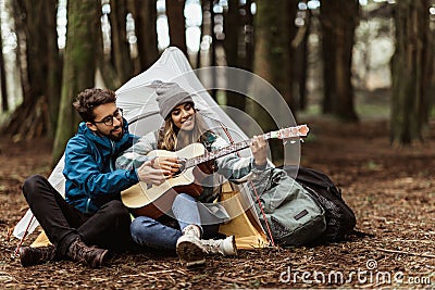 Happy young caucasian guy in jacket give lesson playing guitar to wife, sit near tent, enjoy music, spare time Stock Photo