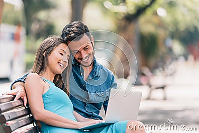 Happy young couple using laptop computer sitting on a bench in city outdoor - Two lovers having fun spending time together Stock Photo