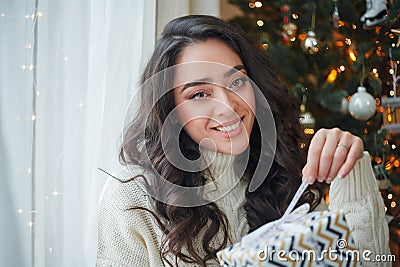 Happy young dark-haired curly woman in a light knitted sweater prepares gifts near the Christmas tree. Stylish model makes online Stock Photo