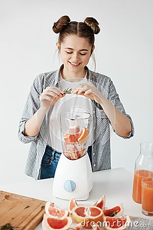 Happy young girl smiling blending healthy detox fresh grapefruit smoothie adding rosemary over white wall. Stock Photo