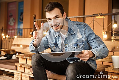 Happy young guy is looking at the camera and showing a victory gesture with his right hand while holding his tablet with his left Stock Photo