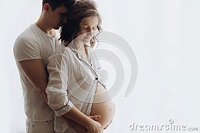 Happy young husband hugging his pregnant wife and holding belly bump. Stylish pregnant family, mom and dad in white pajamas, Stock Photo