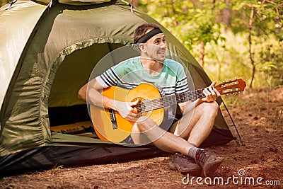 Happy young man camping and strum a guitar instrumental music to relax against background of forest sunset. Stock Photo