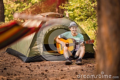 Happy young man camping and strum a guitar instrumental music to relax against background of forest sunset. Wilderness Travel Stock Photo