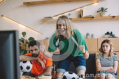 happy young man in green fan t-shirt celebrating while his upset friends sitting behind on sofa during watch of soccer match Stock Photo