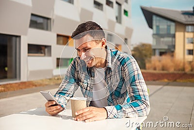 Happy young man sitting outdoors looking in his phone and holding cup of coffee Stock Photo