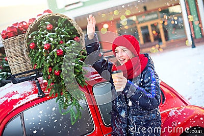 Happy young model girl with hot beverage in hands waves to someone comming. Girl stay near fashion red car ready for shoping Stock Photo